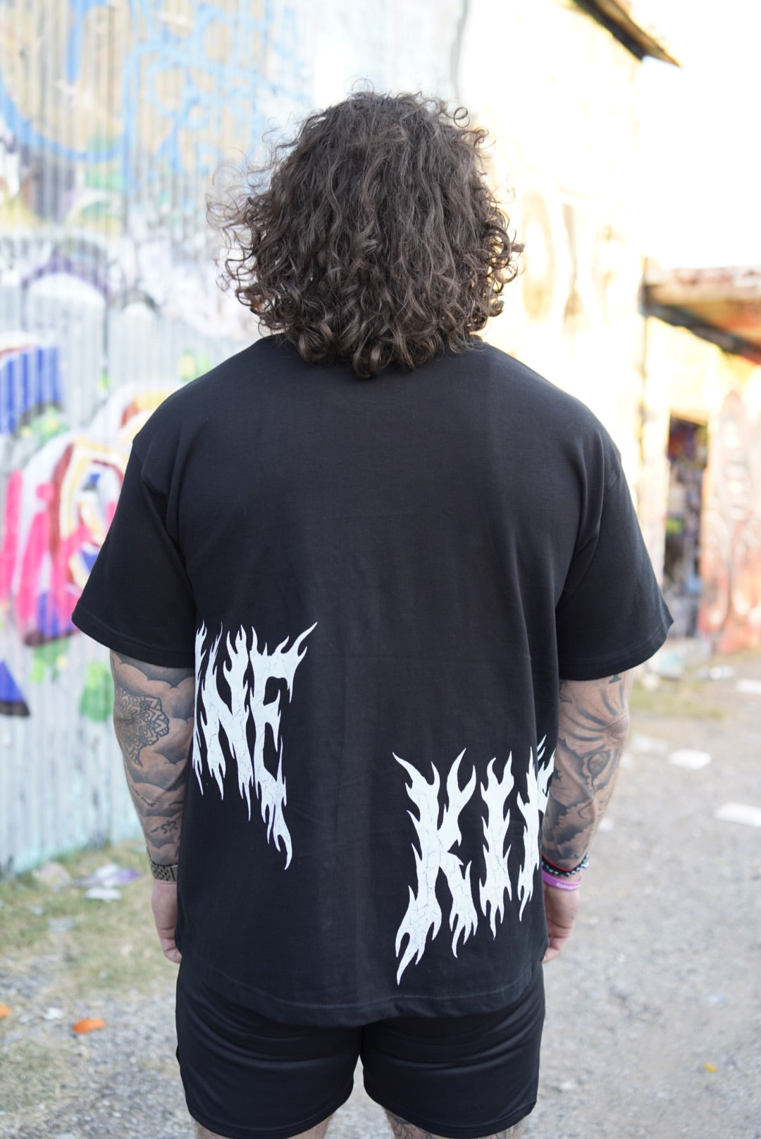 Back view of a man wearing a Black T-shirt with white flamed lettering on the sides reading “KIN” and “SANE”