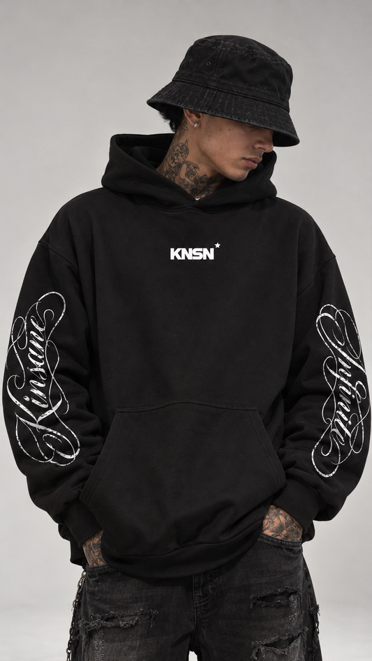 Man wearing Black hoodie with white cursive texts on the sleeves reading “Kinsane” and “Infinite” and small text on the front reading “KNSN*”