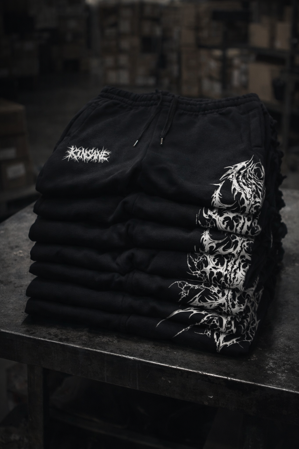 Stack of a few kinsane black cuffed joggers with white text logo reading “KINSANE”  and white tribal designs on a metal surface 