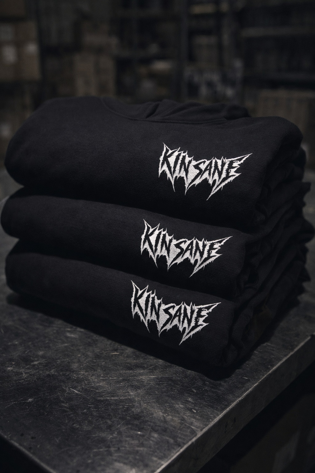 Three black hoodies with white outlined black lettering reading “KINSANE” stacked on a metal surface 