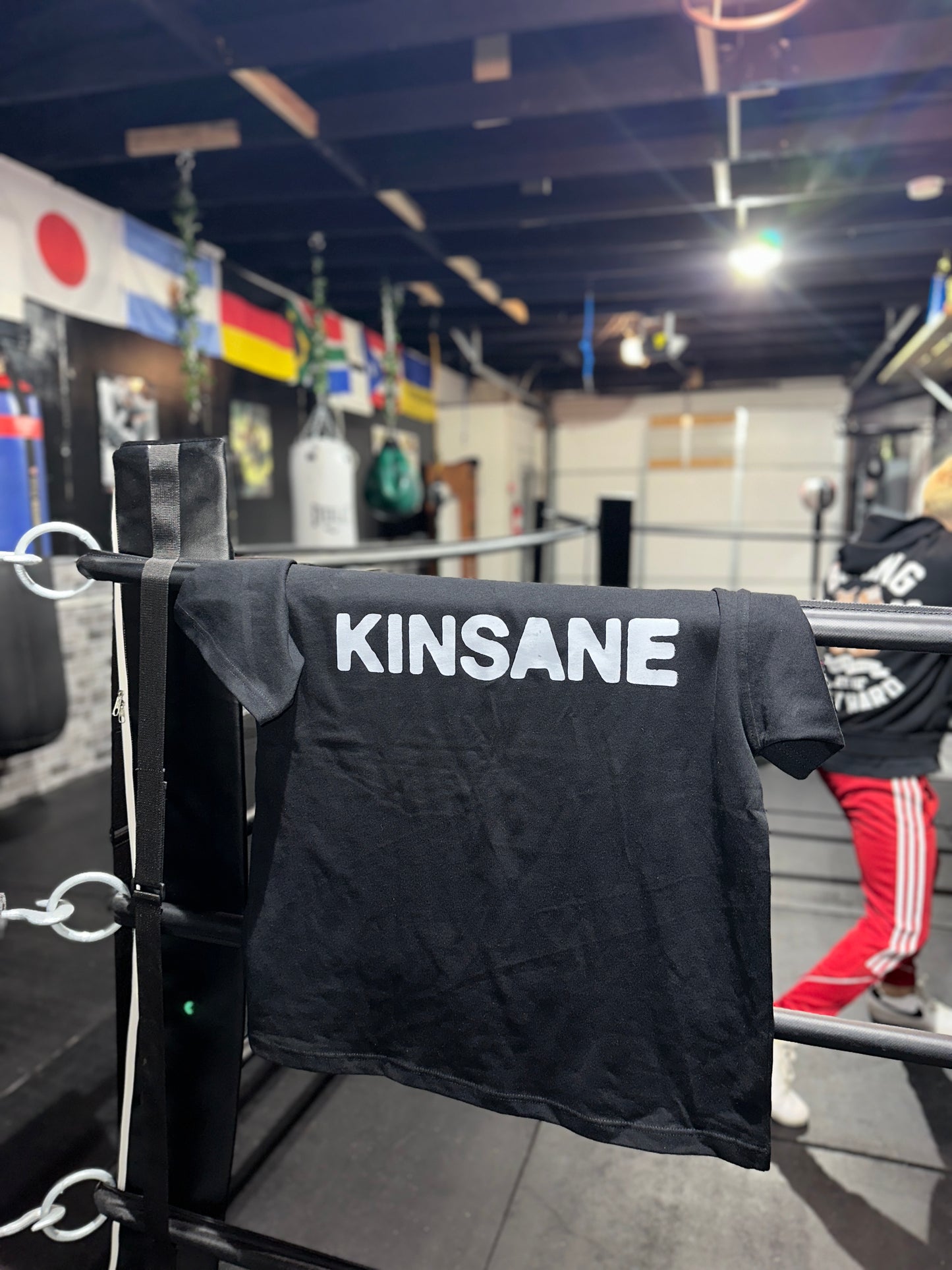 Back view of a black T-shirt with white text reading “KINSANE” Laying on the ropes of a boxing ring in a boxing gym 