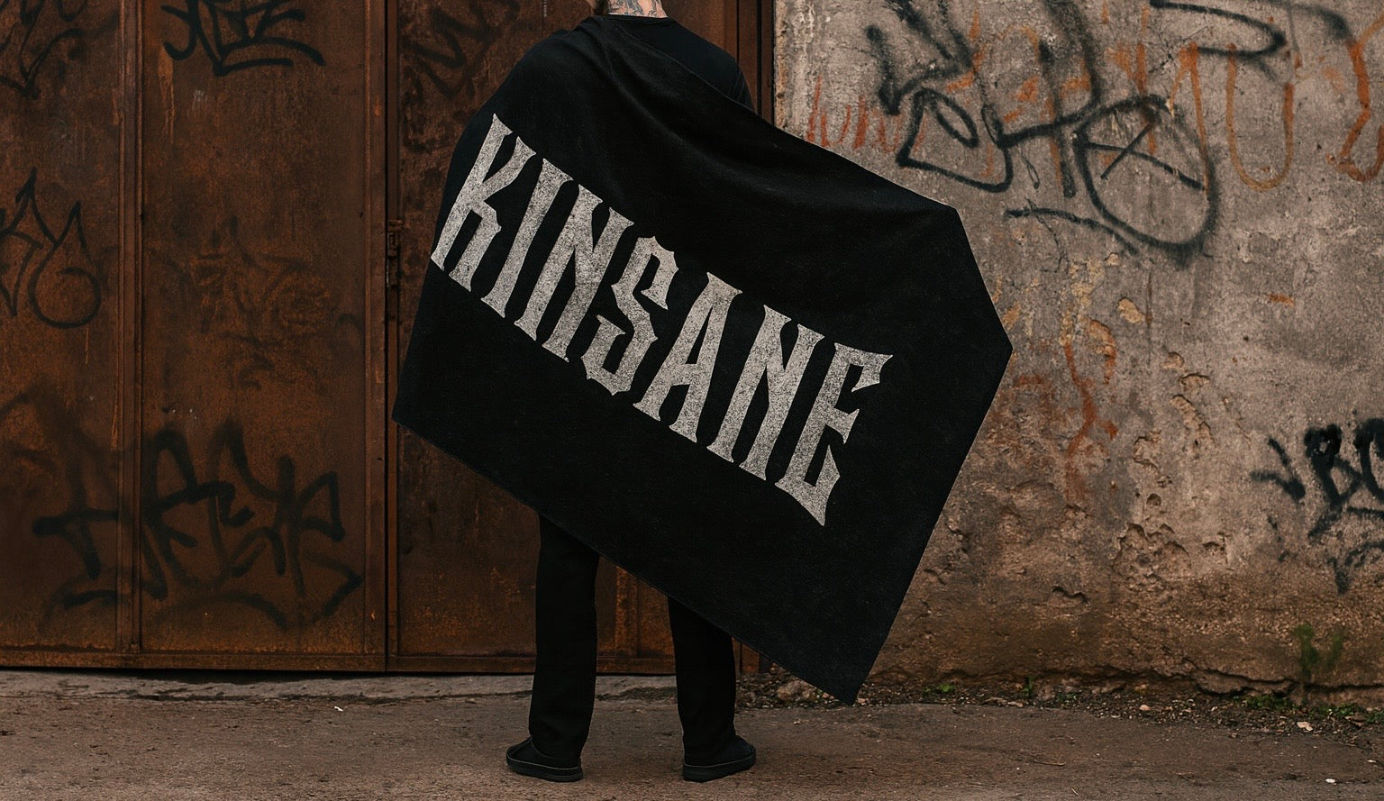 Back view of a man holding a black flag on his back with white lettering reading “KINSANE” 