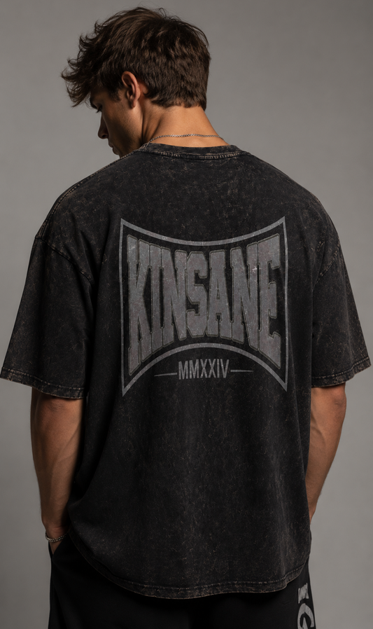 Back view of a man wearing a acid washed black t shirt with grey logo and text reading “KINSANE MMXXIV” 