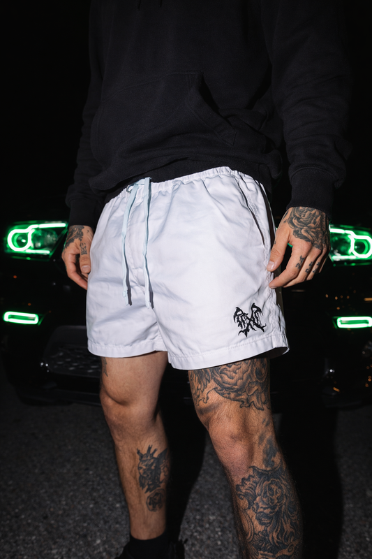 Man standing in a parking lot with a black dodge durango in the background wearing white shorts with white drawstrings with a black logo on the left leg 