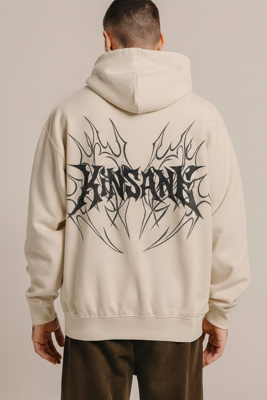 Back view of a man wearing a beige hoodie with black tribal designs and black lettering reading “KINSANE” 