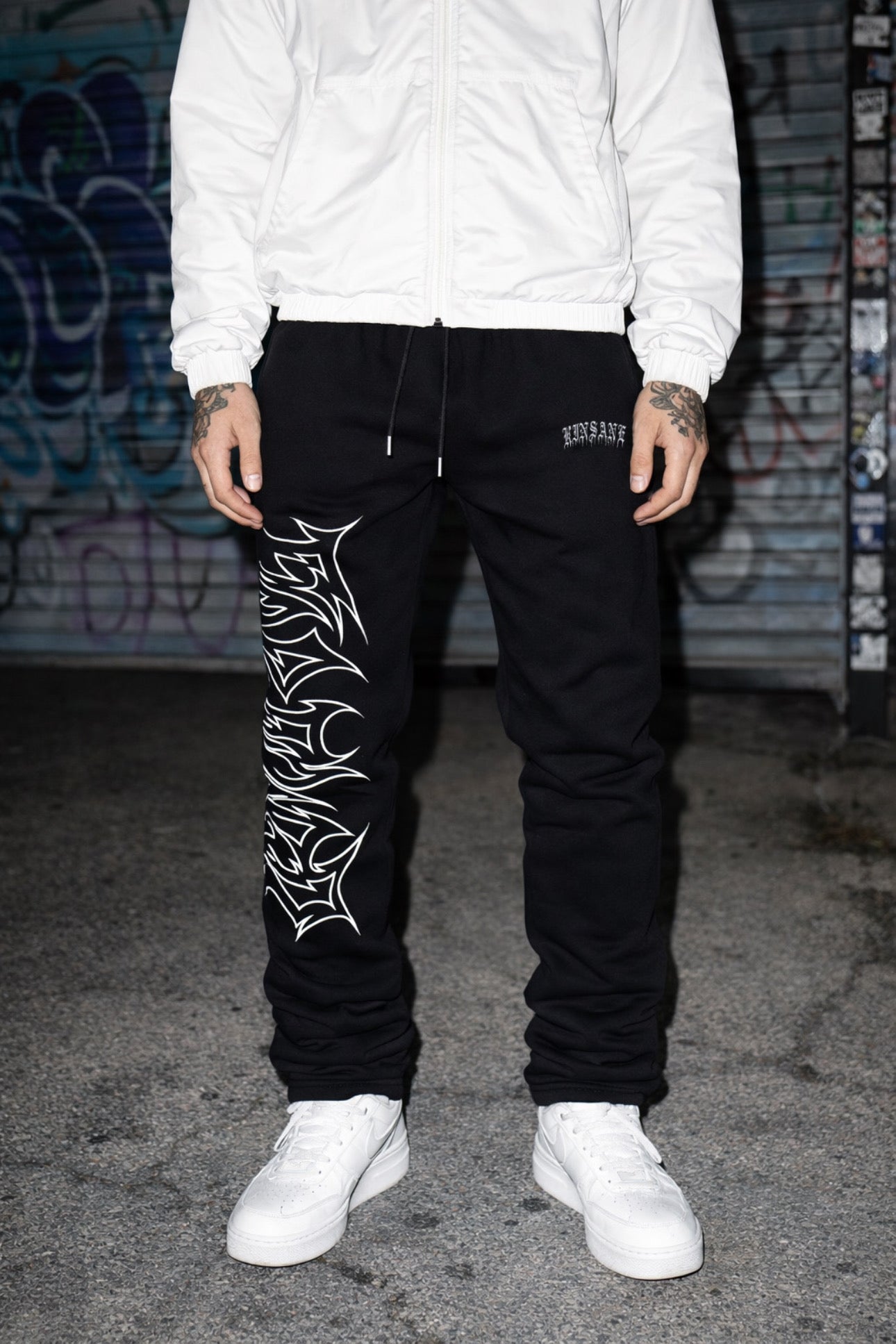 Man wearing black stacked sweatpants with white outlined lettering reading “FOREVER” on the right leg and white lettering reading “KINSANE” on the left leg