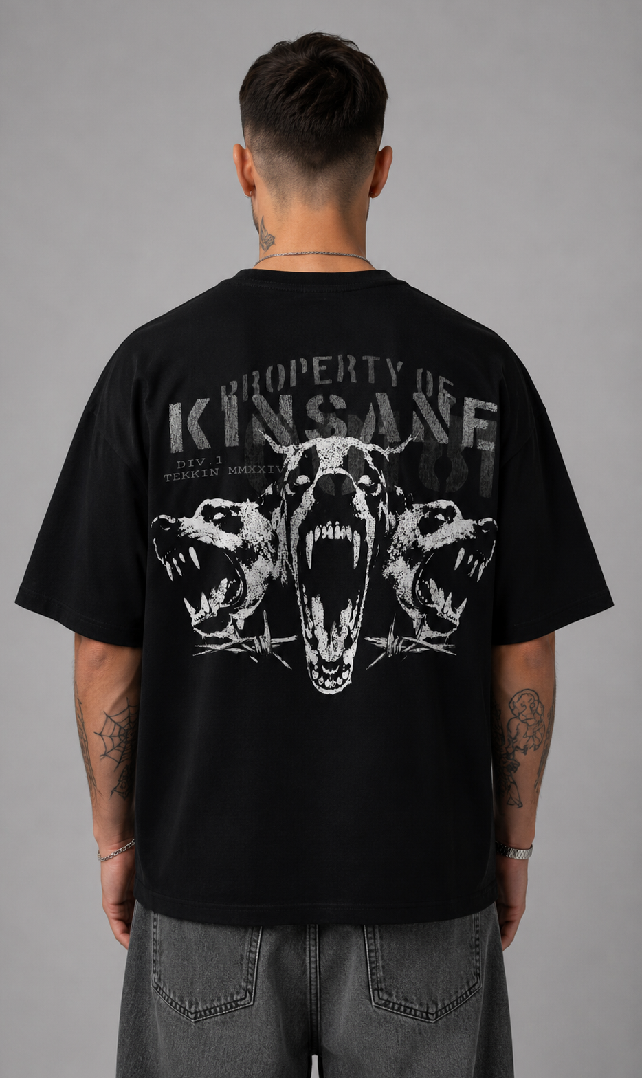 Back view of a man wearing a black t shirt with a white three dogs design and gray text reading “PROPERTY OF KINSANE DIV. 1 TEKKIN MMXXIV” 