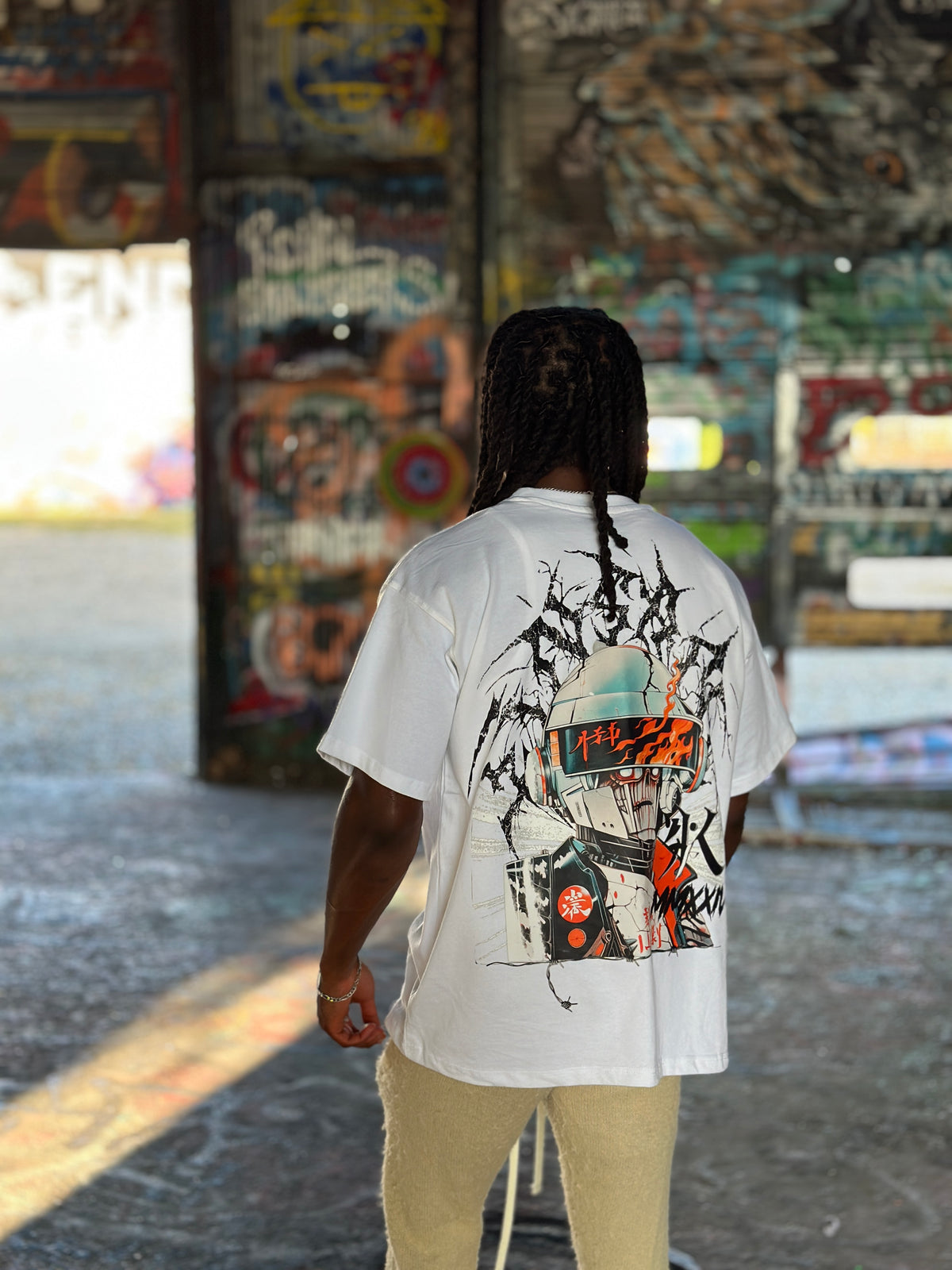 Back view of a man wearing a white t shirt with a multi color design of a person with helmet gear on with cracks and flames on it and black lettering behind it reading “KINSANE” and other black lettering reading “MMXXIV” and a black K logo with barbed wire at the bottom 
