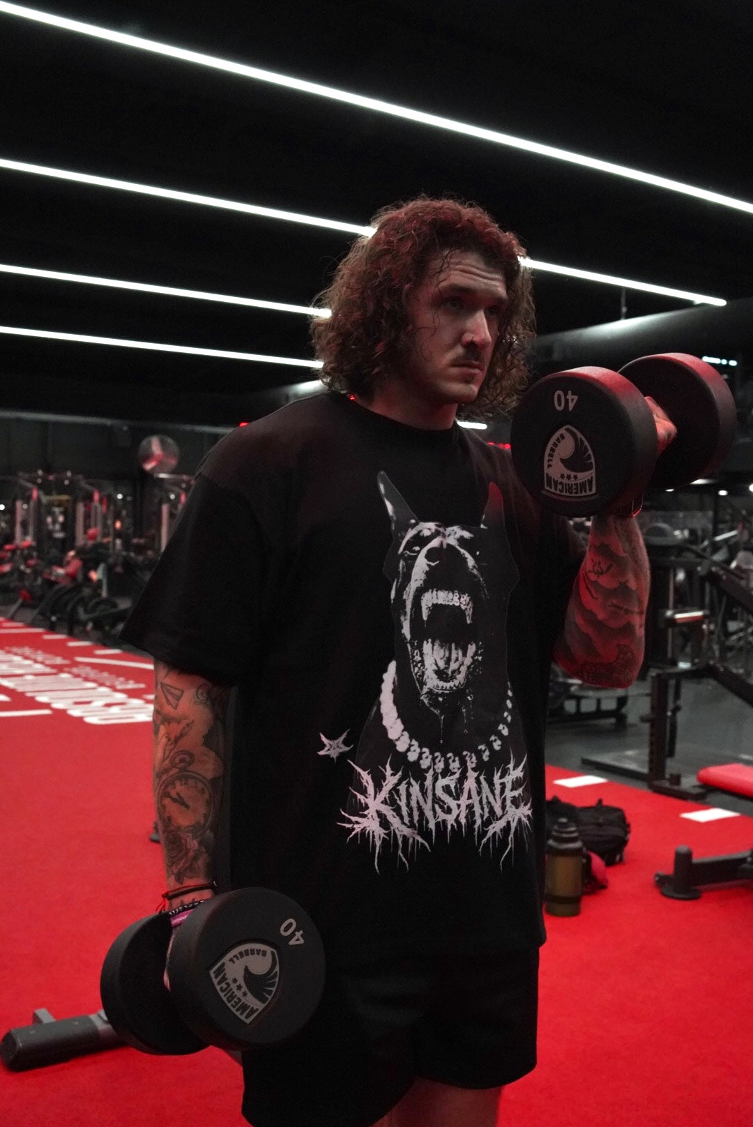 Man lifting weights wearing a Black T-shirt with a white and black Doberman dog design and white tribal lettering reading “KINSANE” 