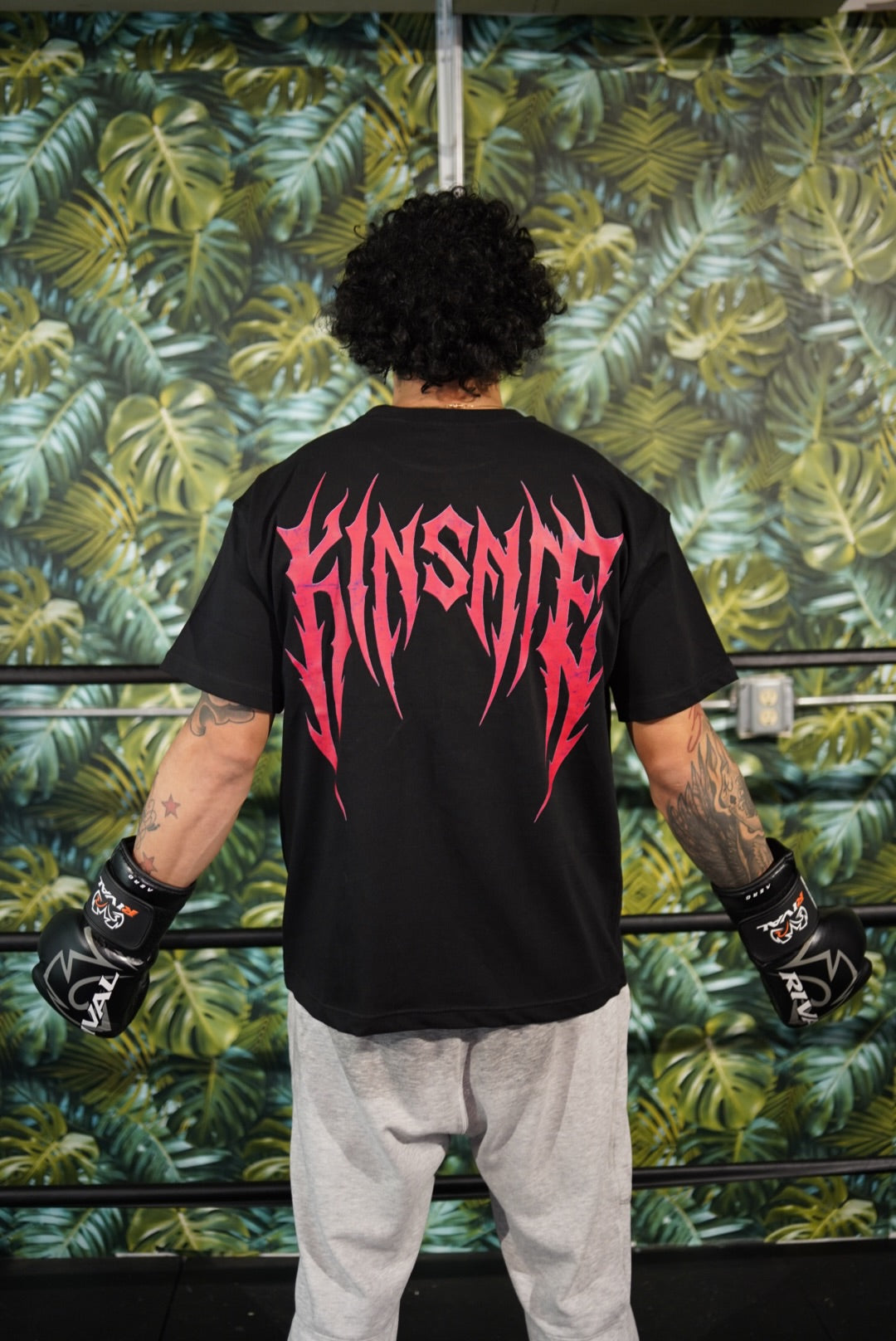 Back view of a man wearing a black T-shirt with red tribal lettering reading “KINSANE” 