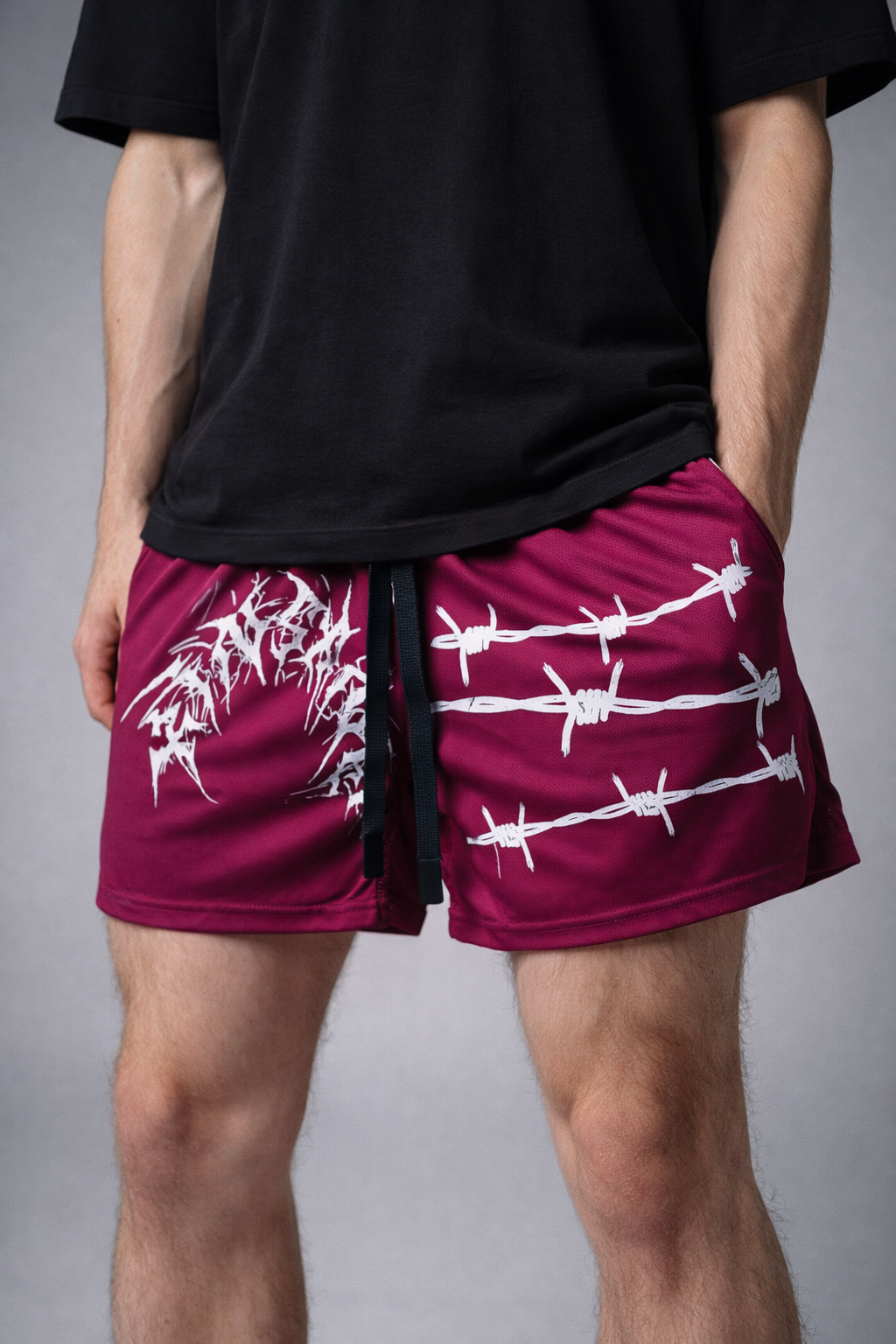 Man wearing Dark red athletic shorts with elastic waistband and black drawstrings and white tribal lettering reading “KINSANE” on the right leg and white barbed wire design on the left leg 