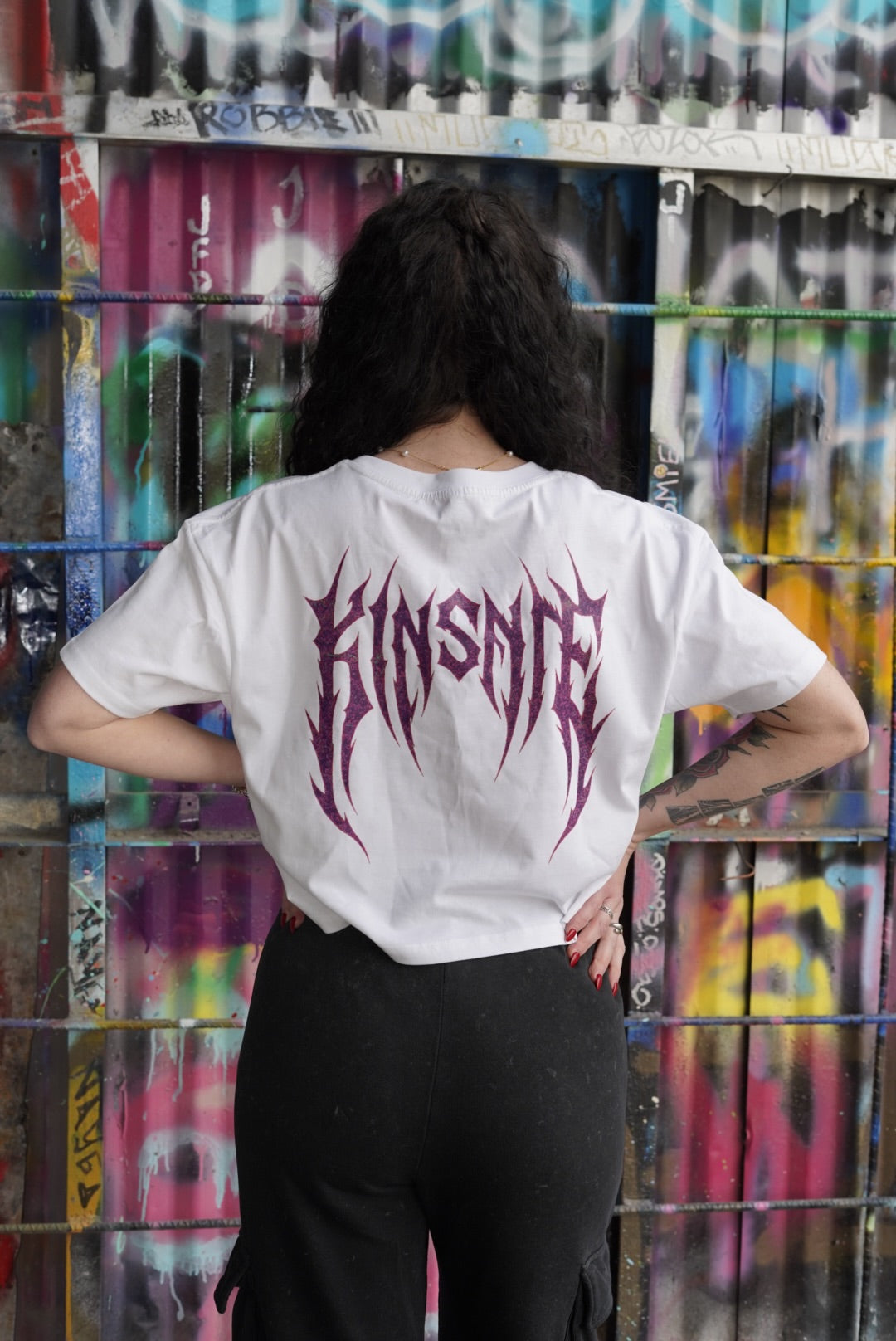 Back view of a girl wearing a white crop top T-shirt with dark purple tribal lettering reading “KINSANE” 
