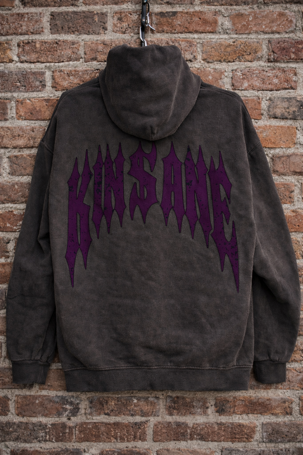 Back view of a vintage gray hoodie with dark purple text reading “KINSANE” Hanging in front of a brick wall 
