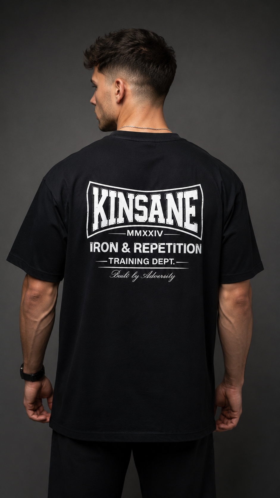 Back view of a man wearing a black t shirt with white logo and white text reading “KINSANE MMXXIV IRON & REPETITION TRAINING DEPT. BUILT BY ADVERSITY” 