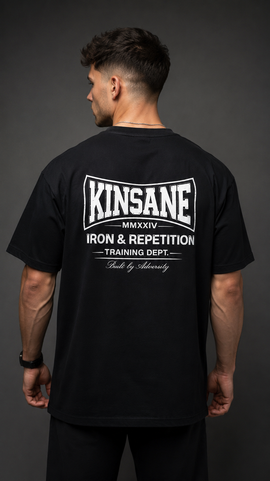 Back view of a man wearing a black t shirt with white logo and white text reading “KINSANE MMXXIV IRON & REPETITION TRAINING DEPT. BUILT BY ADVERSITY” 