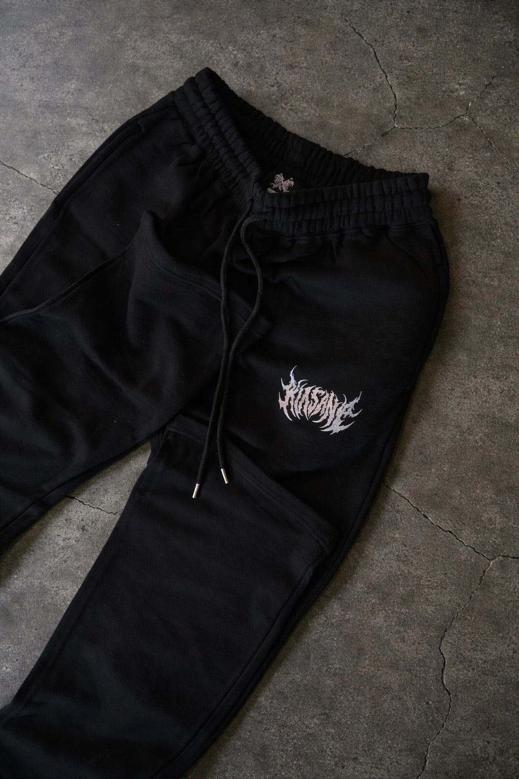 Black stacked sweatpants with white lettering reading “KINSANE” on the left leg laying on concrete 