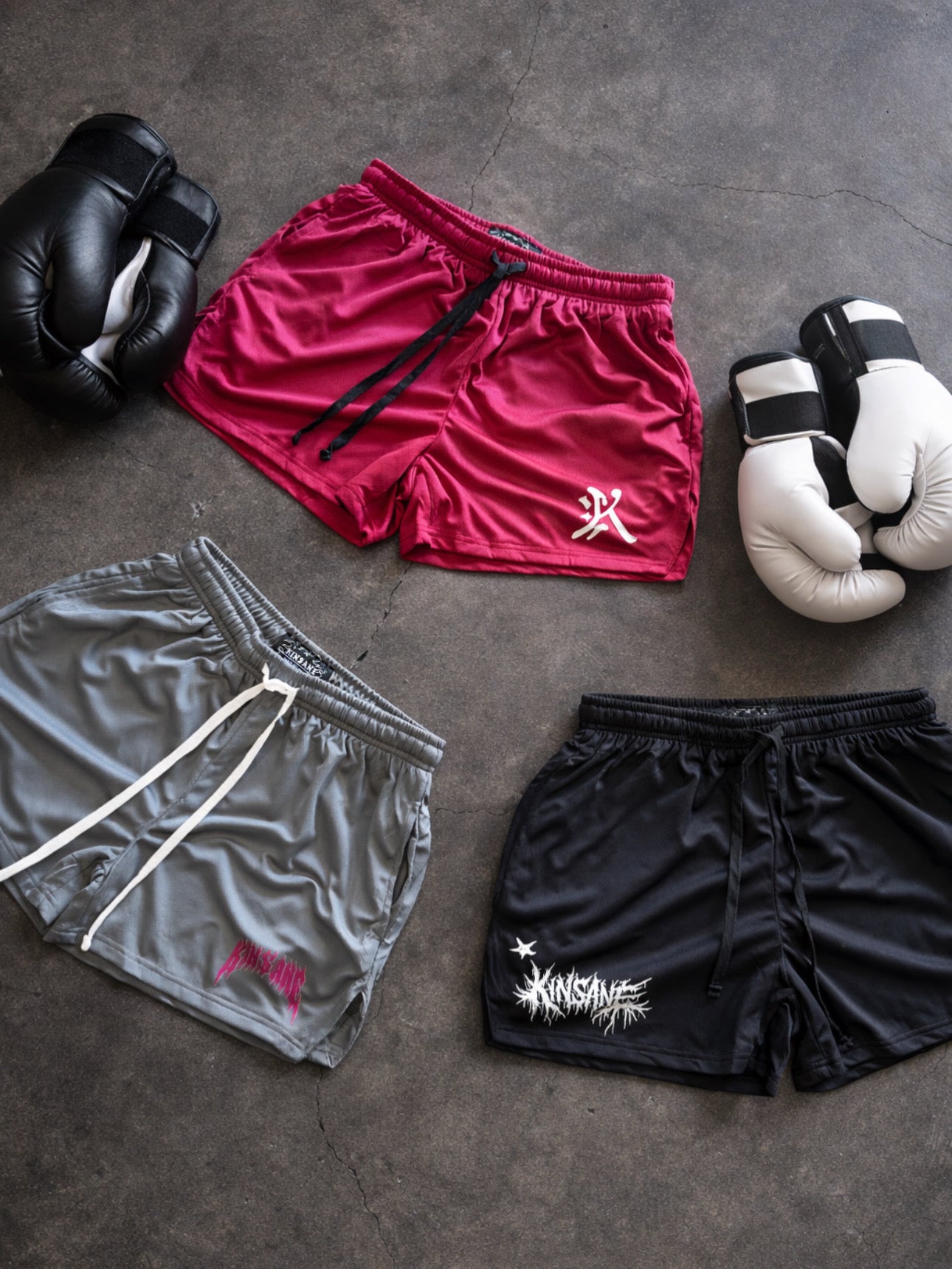3 Pairs of shorts laying on the ground next to two pairs of boxing gloves and the shorts are grey shorts with white drawstrings and a pink logo reading “Kinsane” on the left leg, black athletic shorts with black drawstrings with white lettering reading “KINSANE” on the right leg, and red athletic shorts with black drawstrings with a silver logo of the letter “K” on the left leg. 
