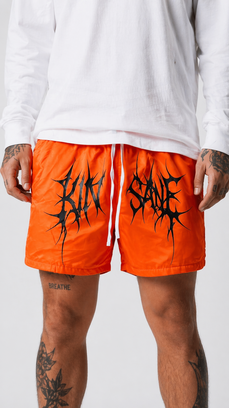 Man wearing Orange trunks with white drawstrings and black tribal lettering across the middle reading “KINSANE” 