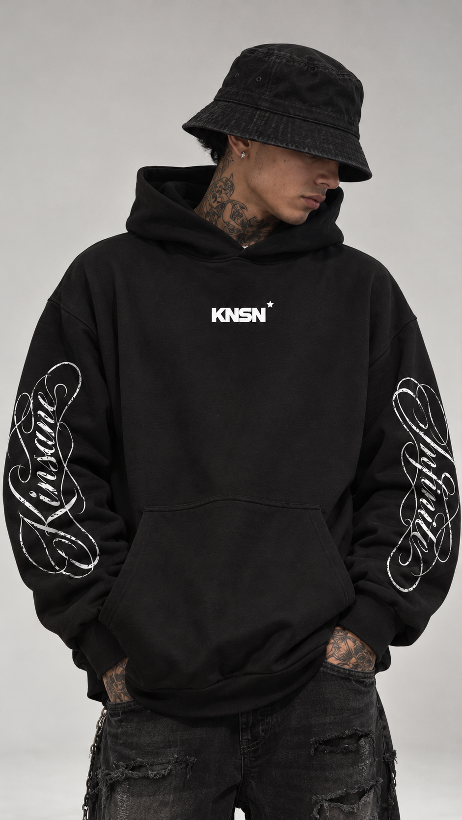Man wearing Black hoodie with white cursive texts on the sleeves reading “Kinsane” and “Infinite” and small text on the front reading “KNSN*”