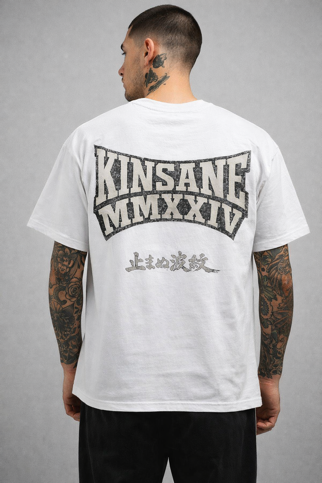 Back view of a man wearing a white T-shirt with silver lettering reading “KINSANE MMXXIV” and silver Japanese lettering translating to “THE RIPPLE THAT CAN NOT BE STOPPED” 