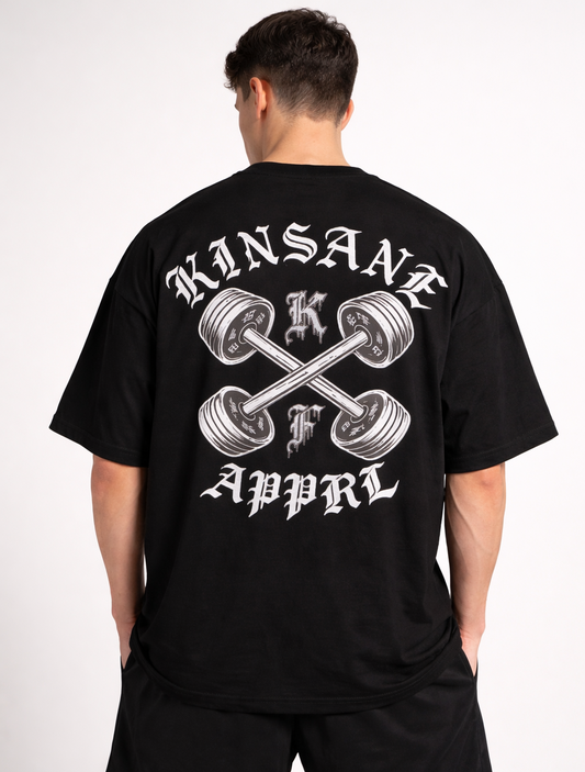 Back view of a man wearing a black t shirt with a white weights crossing logo with white lettering reading “KINSANE APPRL” and silver lettering single letters “K” and “F”